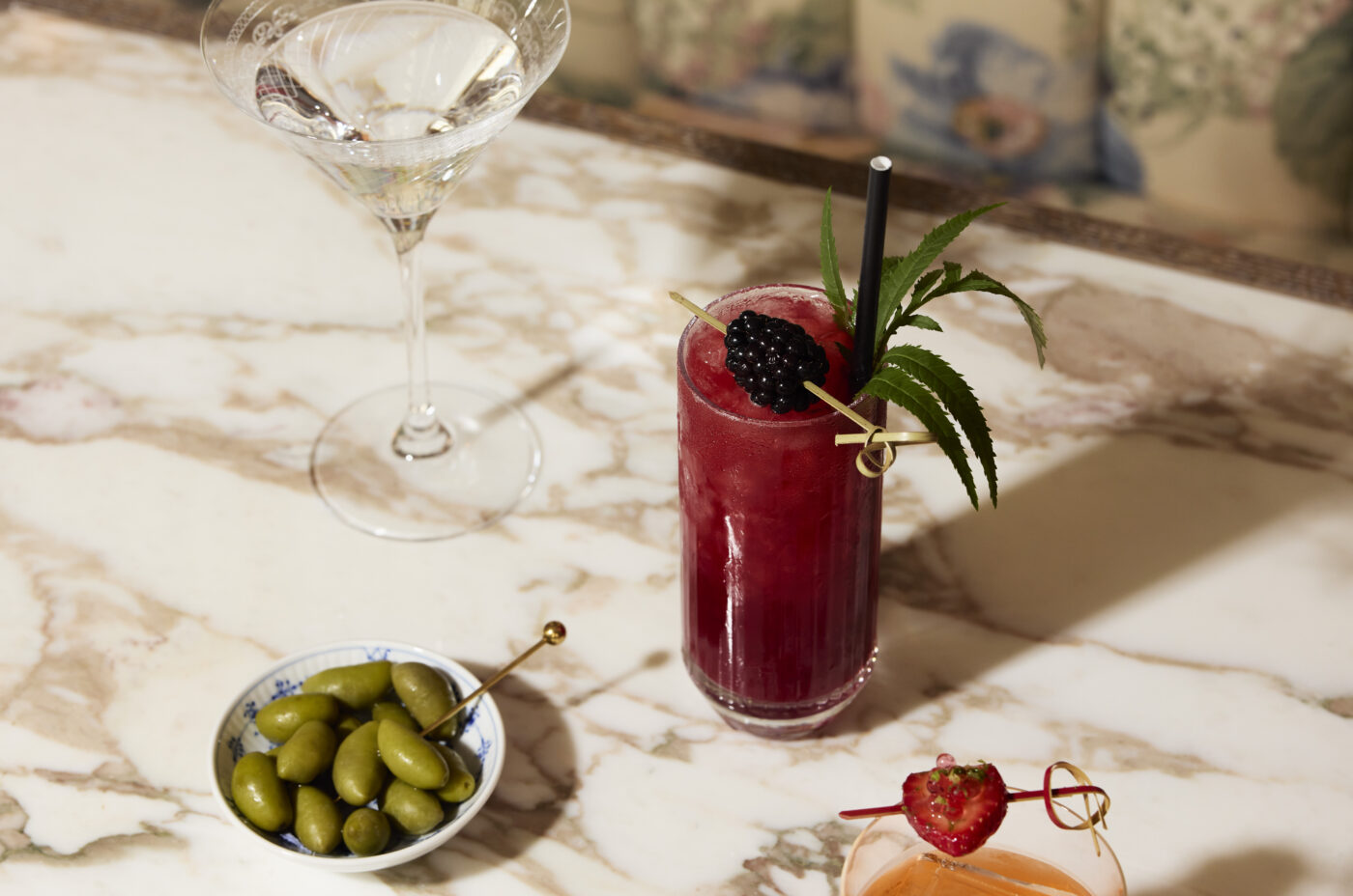 A classic Martini and two cocktails garnished with fruit sit alongside a bowl of green olives on a marble table, in front of a floral banquette.
